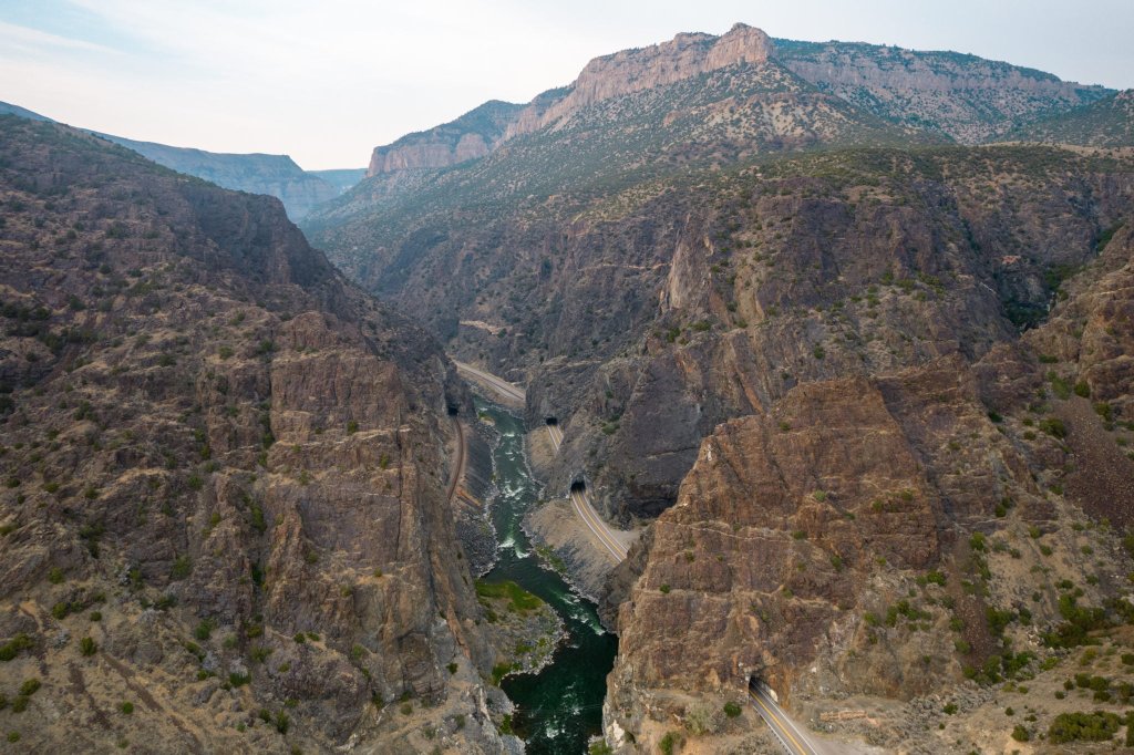 A vast canyon with a winding river and towering rocky cliffs, featuring a highway cutting through the mountains in the distance in Wind River Country.