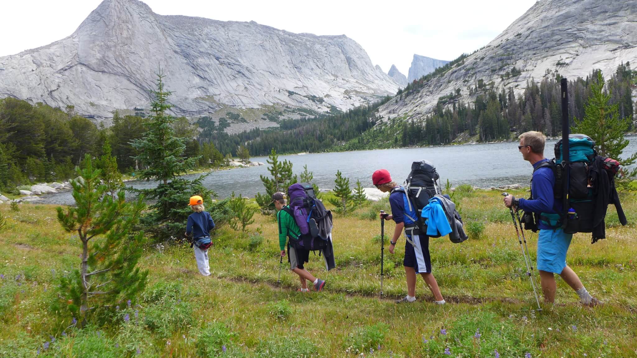 A group of hikers with backpacks and trekking poles walk along a mountain trail near a serene lake, surrounded by towering peaks and lush greenery in Wind River Country.