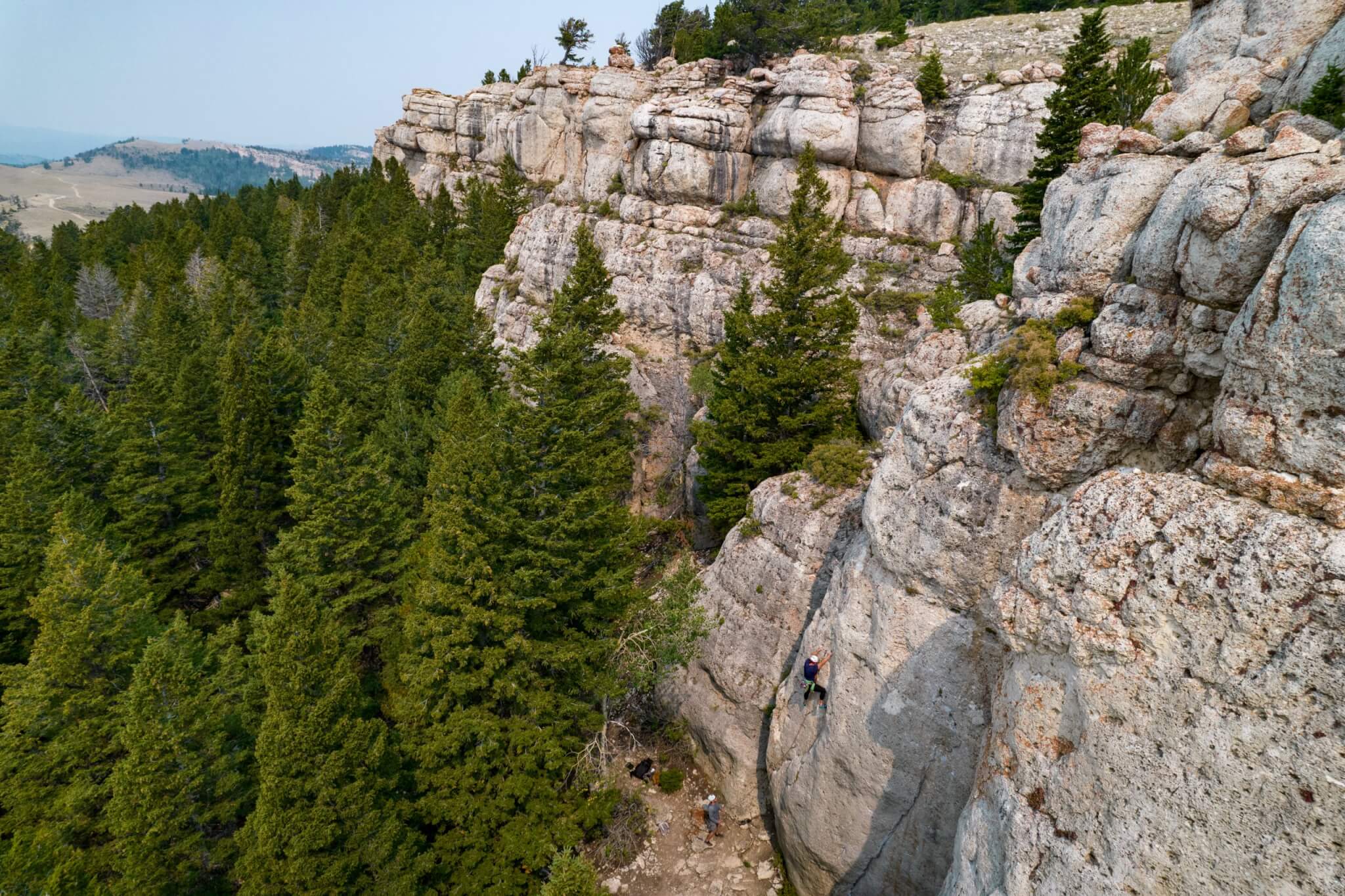 A climber ascends a steep rock face on a rugged mountain ridge, surrounded by dense evergreen forests and distant rolling hills in Wind River Country.