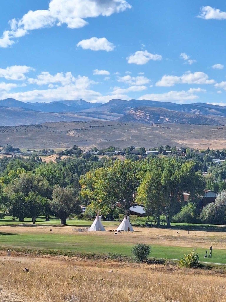 A serene landscape featuring two white teepee tents set up in a grassy field with a backdrop of lush trees and distant mountains under a clear blue sky with scattered clouds in Wind River Country.