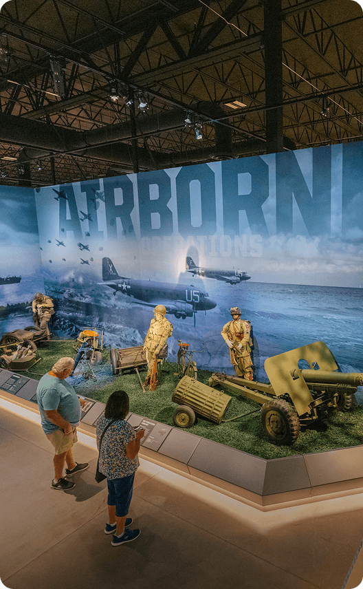 Visitors explore a World War II-themed exhibit featuring life-size dioramas, military vehicles, and aircraft silhouettes against a backdrop of a dramatic sky in Wind River Country.