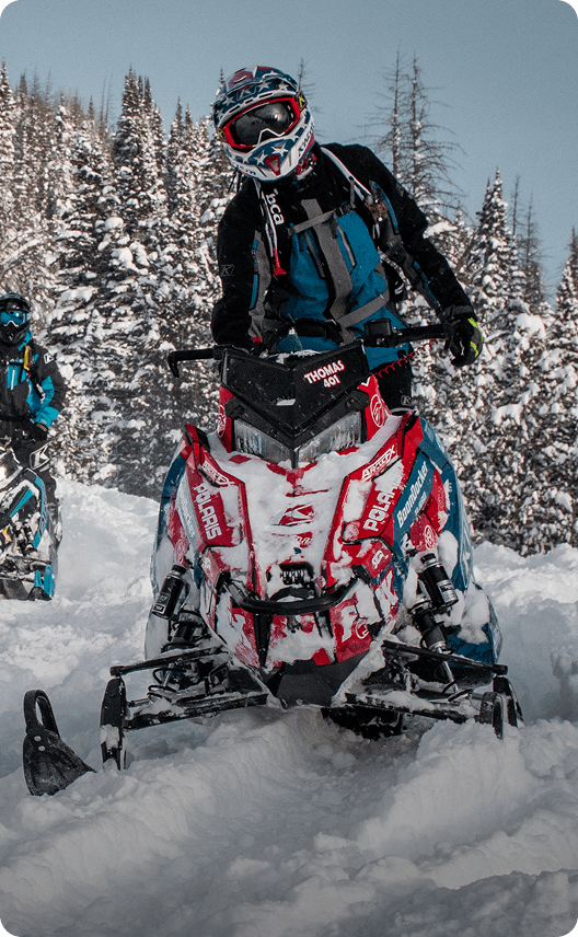 A snowmobiler in a blue and black jacket rides through a snowy forest on a red and black snowmobile in Wind River Country.