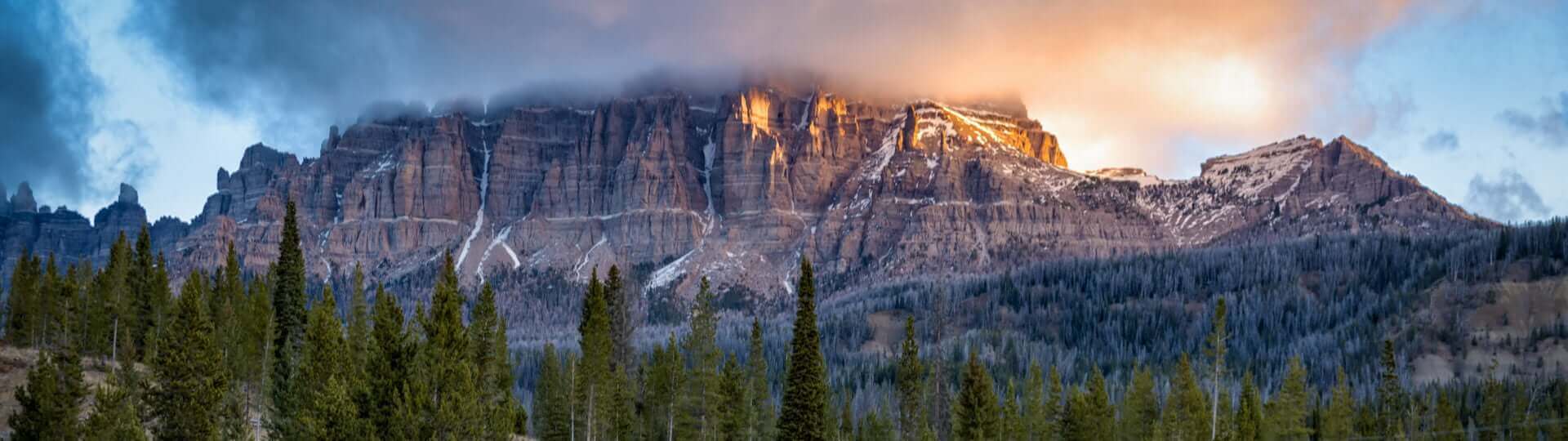 A majestic mountain range with snow-capped peaks rises above a dense forest of evergreen trees, bathed in the warm glow of a sunrise or sunset in Wind River Country.
