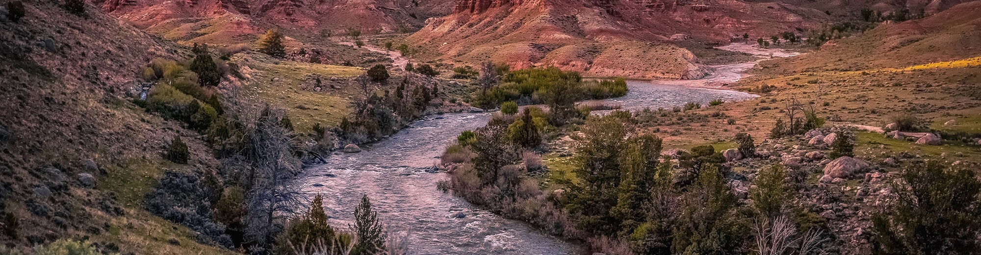 A panoramic view of a winding river flowing through a rugged, colorful canyon with towering red rock formations and lush greenery along its banks in Wind River Country.