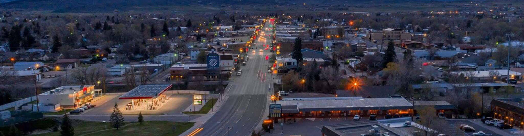 A panoramic view of a small town at dusk, with a main street lined with illuminated buildings and a gas station on the outskirts in Wind River Country.