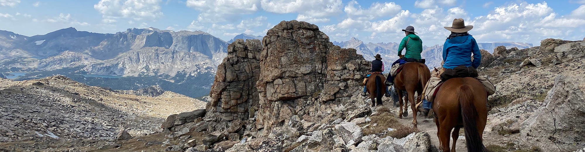 Four riders on horseback traverse a rugged mountain trail with towering peaks and a vast valley in the distance in Wind River Country.