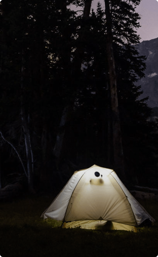 A lone tent illuminated by a lantern in a dark forest clearing at night in Wind River Country.