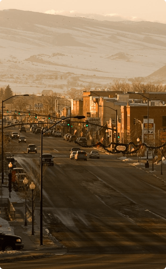 A bustling town street at dusk, lined with snow-covered sidewalks and illuminated by streetlights, with traffic moving along the road in Wind River Country.