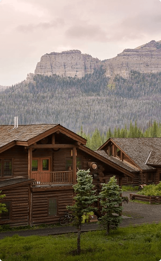 A rustic log cabin nestled among evergreen trees with a majestic mountain range in the background in Wind River Country.