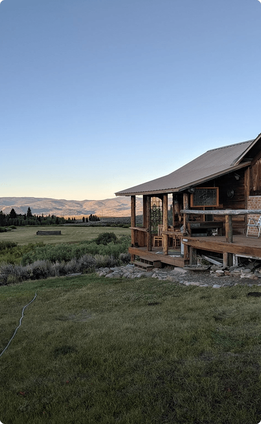 A rustic wooden cabin with a wrap-around porch sits nestled in a lush green meadow, overlooking a breathtaking mountain range at sunset in Wind River Country.