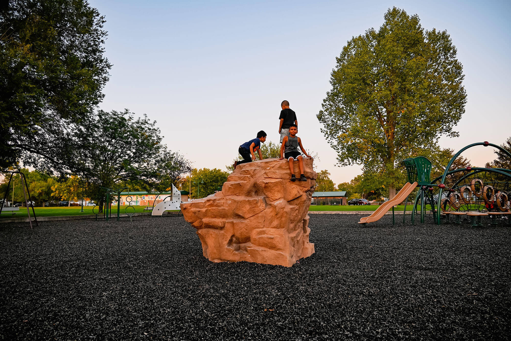 Four children play on a large rock formation at a playground, with one child climbing to the top and the others watching from below in Wind River Country.