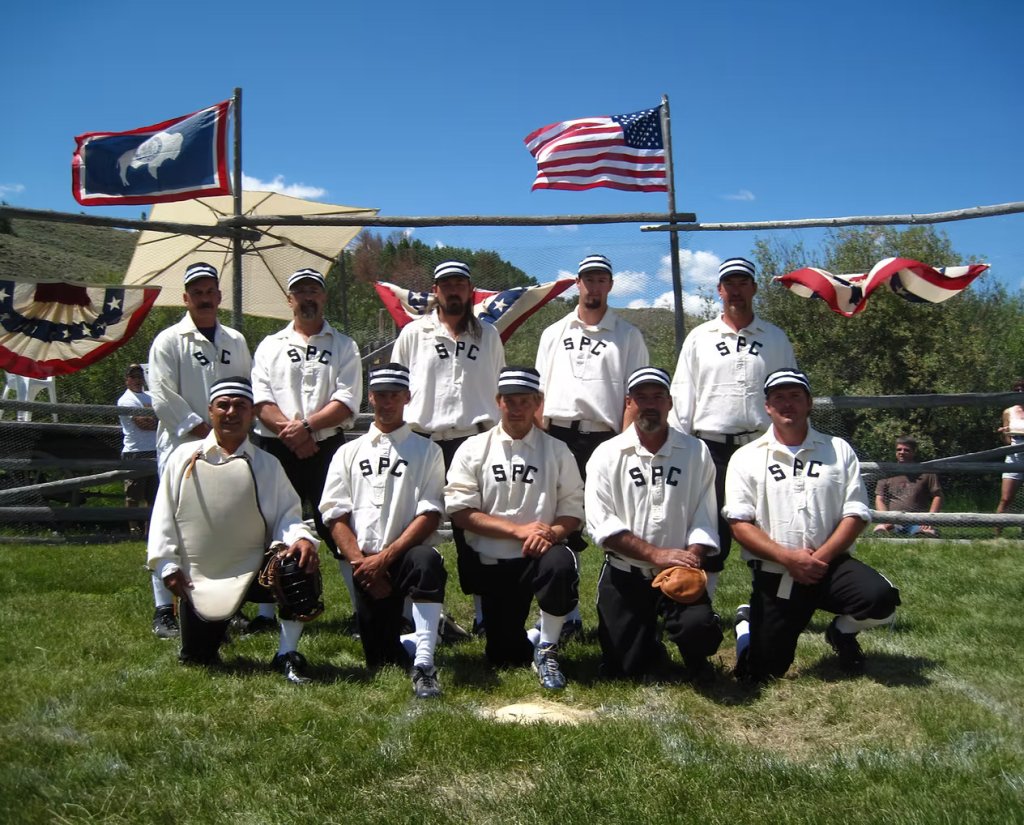A vintage base ball team from South Pass City poses for a group photo on a grassy field, with American and Wyoming state flags in the background during Gold Rush Days in Wind River Country.