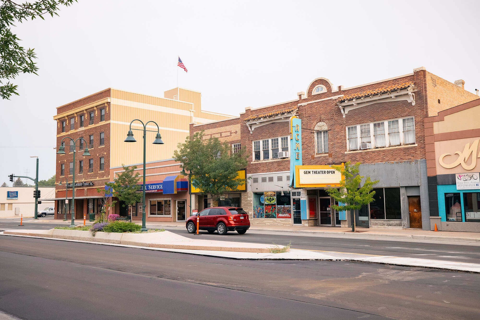 A quaint town square with historic brick buildings, a red car parked on the street, and a movie theater marquee advertising 'Gem Theater Open' in Wind River Country.