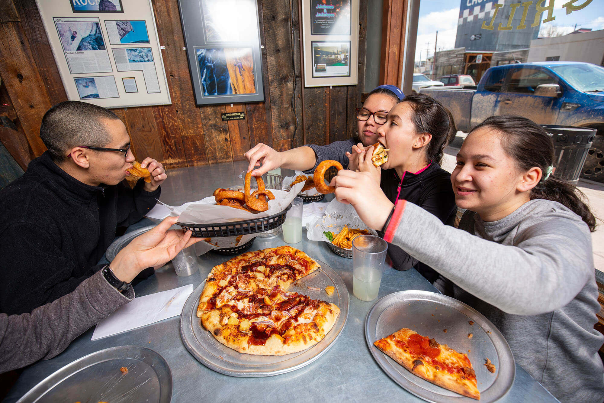 A group of four people enjoying a meal together at a restaurant, with pizza, onion rings, and drinks on the table in Wind River Country.