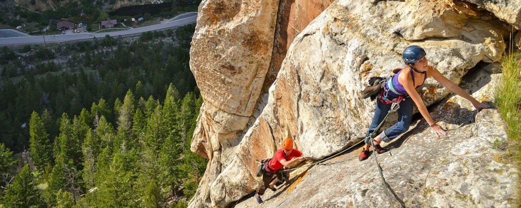 Two climbers ascend a steep rock face, one in a purple tank top and the other in a red shirt, both secured by ropes and wearing helmets, with a lush green forest and winding road visible far below in Wind River Country.
