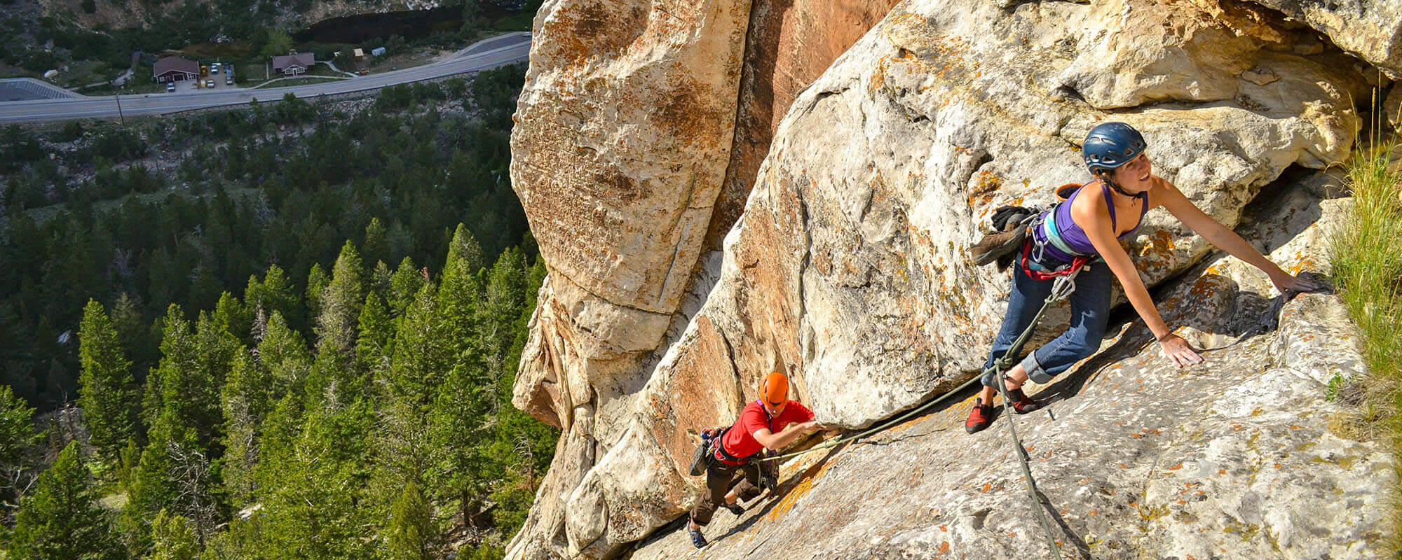 Two climbers ascend a steep rock face, one in a purple tank top and the other in a red shirt, both secured by ropes and wearing helmets, with a lush green forest and winding road visible far below in Wind River Country.