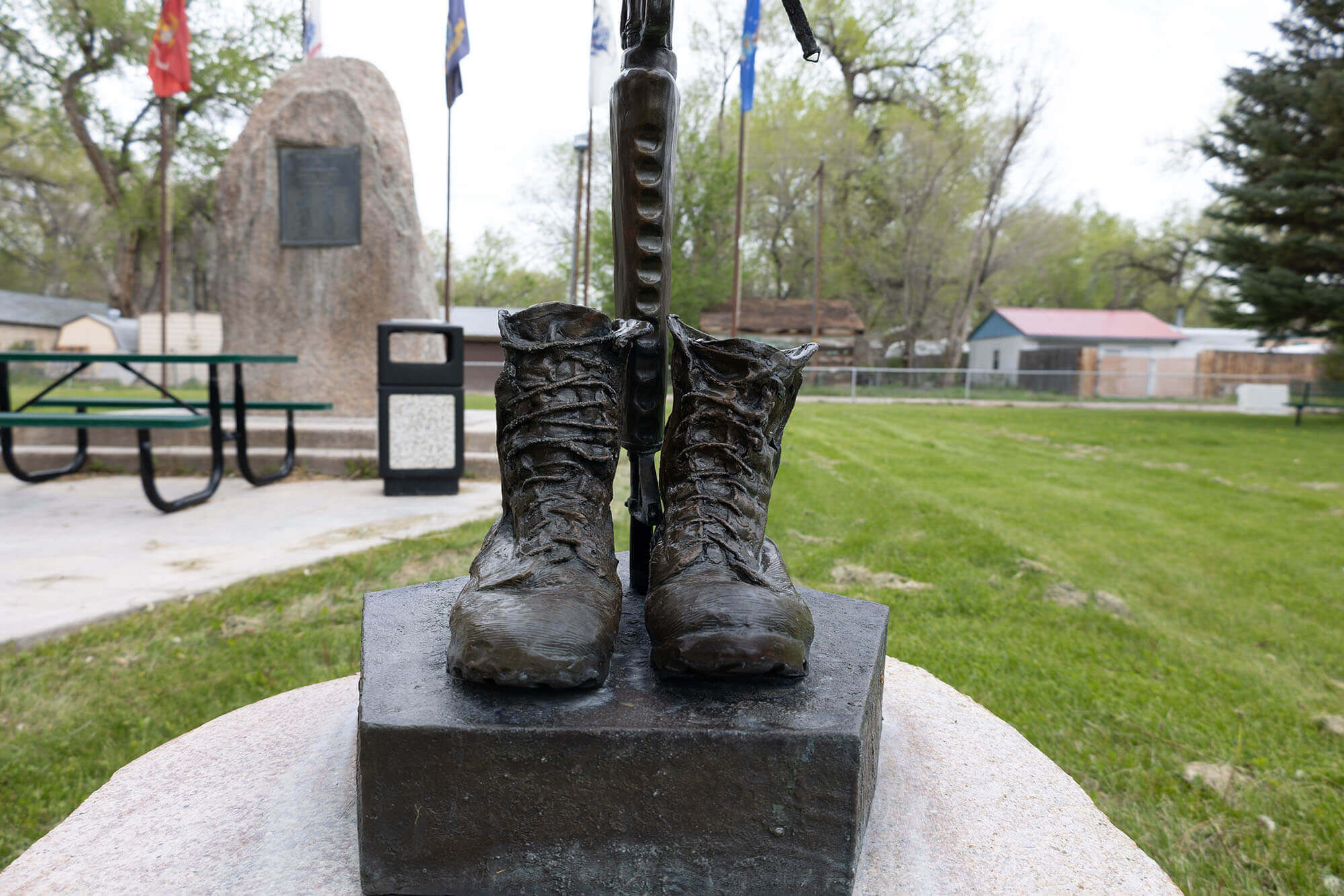 A bronze statue of a pair of military boots stands on a pedestal in a park, surrounded by green grass and trees in Hudson, Wyoming.