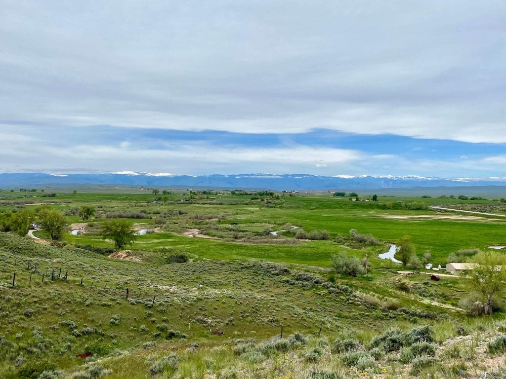 A vast, rolling green landscape with a winding river, dotted with trees and shrubs, stretches towards distant snow-capped mountains under a partly cloudy sky in Hudson, Wyoming.