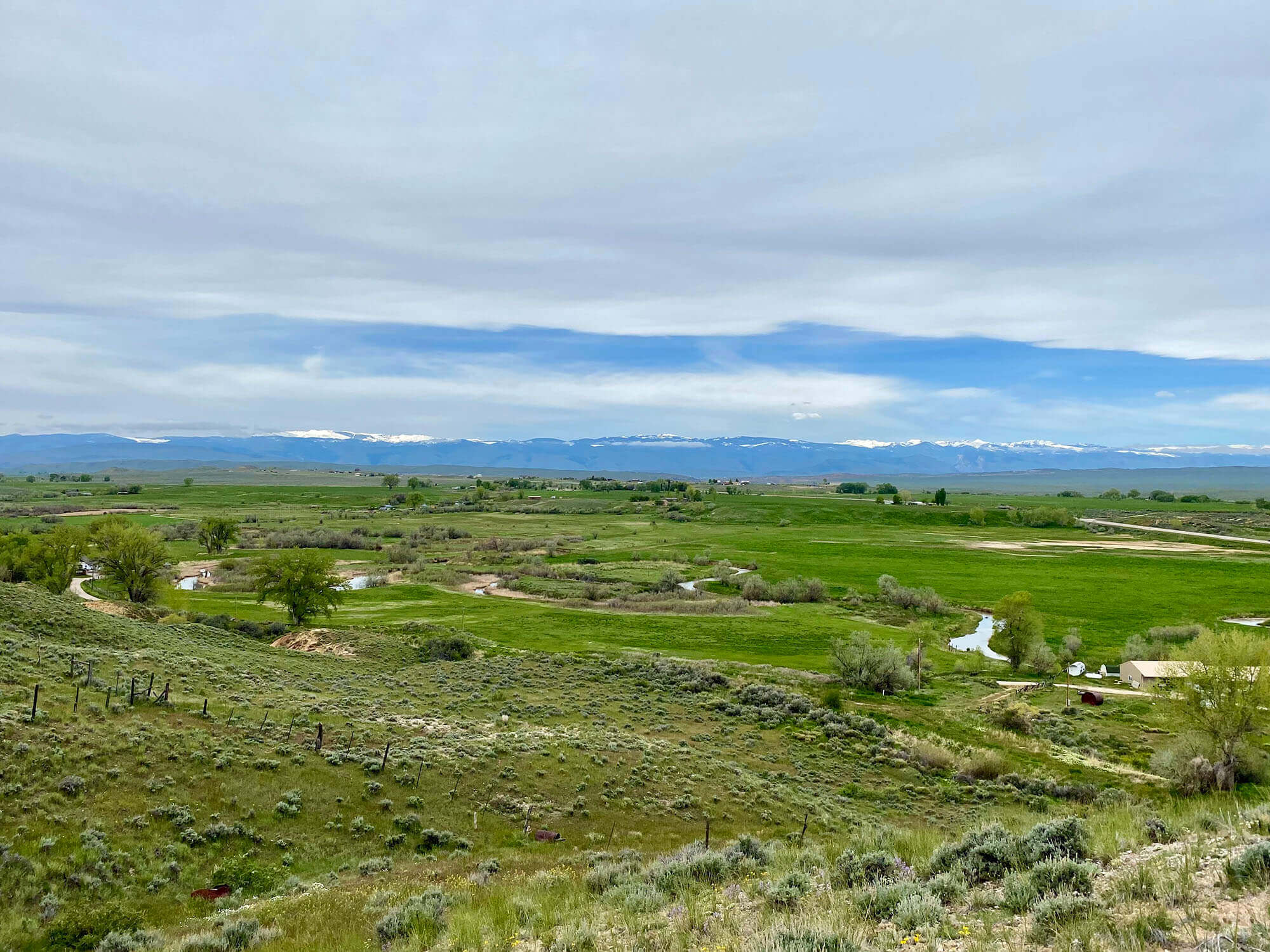 A vast, rolling green landscape with a winding river, dotted with trees and shrubs, stretches towards distant snow-capped mountains under a partly cloudy sky in Hudson, Wyoming.