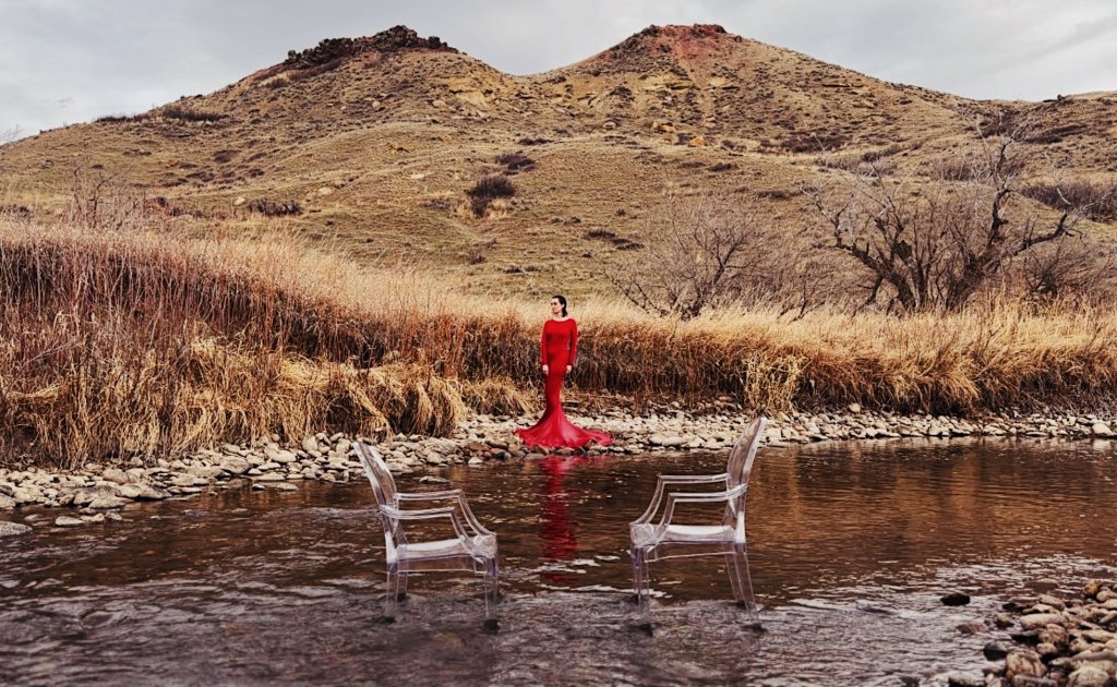 A woman in a red dress stands by a stream with two clear chairs in Wind River Country.